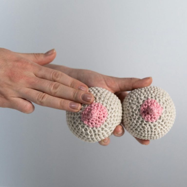 Brustsprechstunde Eine Hand berührt zwei häkelnachgeahmte Brüste / Mamma / Mammae mit rosa Brustwarzen.